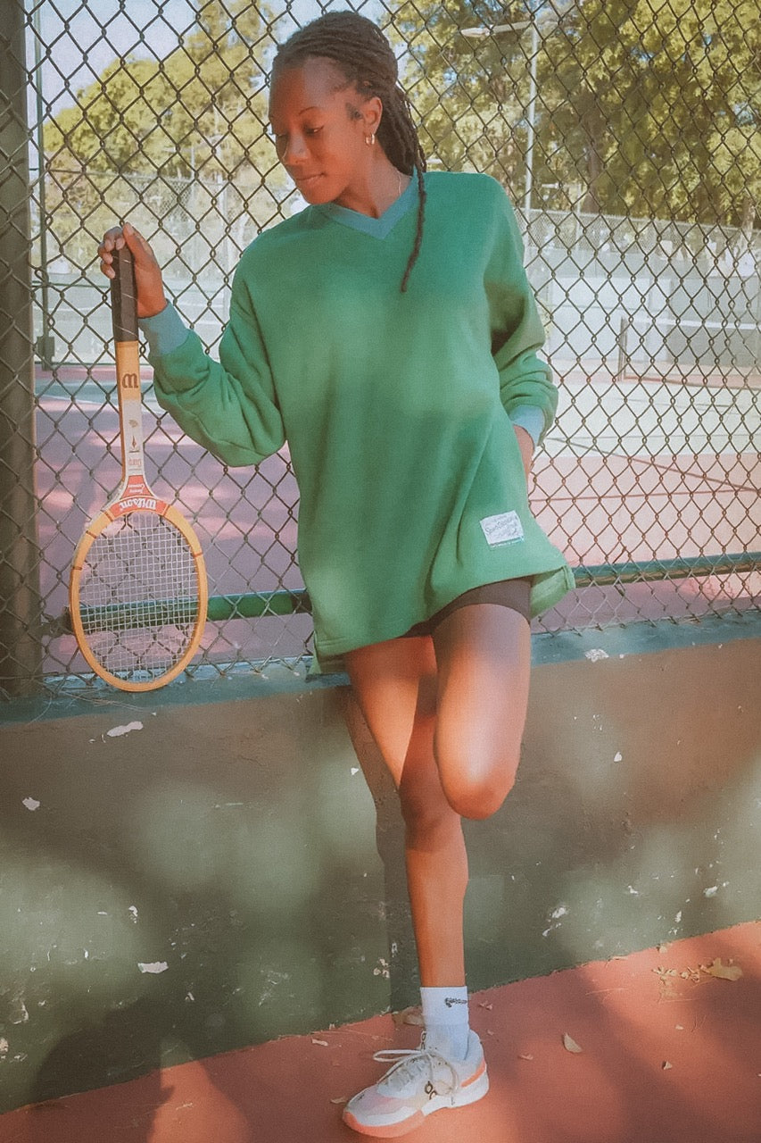 Person in green sweatshirt and shorts holding a tennis racket on a tennis court.