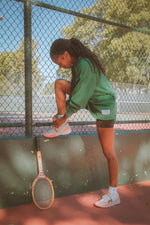 Person on a tennis court with a racket, wearing green shorts and white shoes.