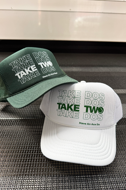 Two tennis hats, one green and one white, with text on a metallic surface.