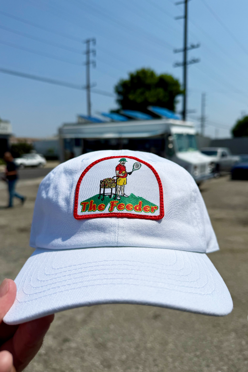 White cap with 'The Feeder' logo held in front of a blurred background