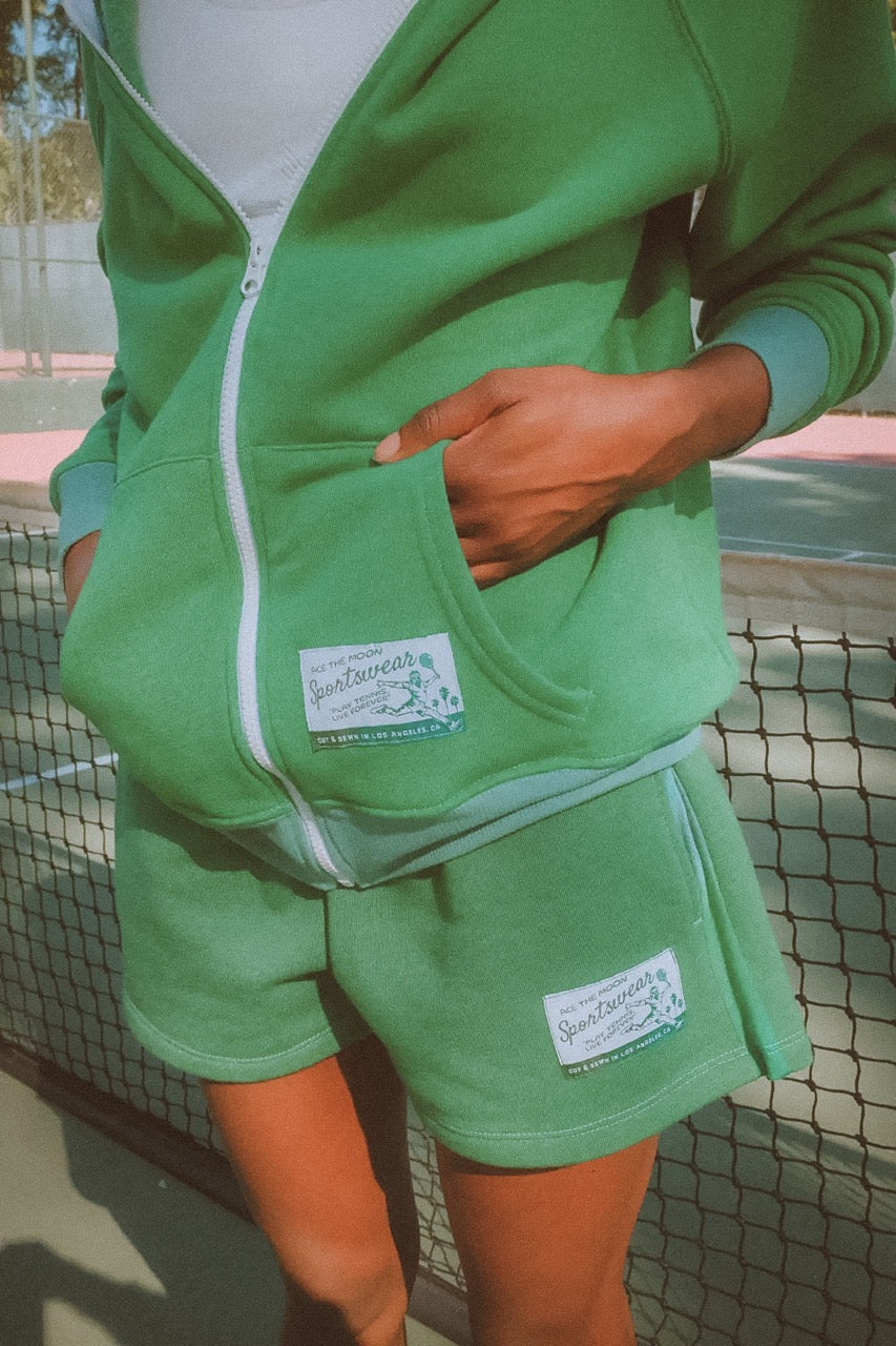 Green outfit with a brand label worn by a person, standing against a blurred background.