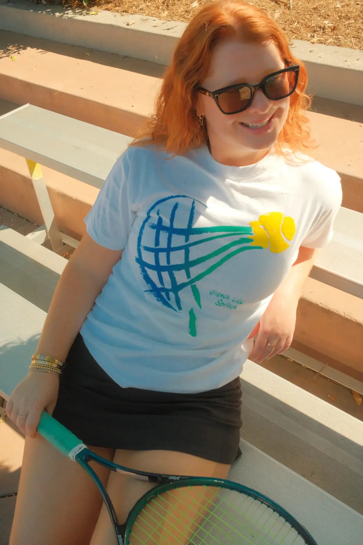 Woman wearing a white t-shirt with a tennis racket design, sitting on a wooden bench.