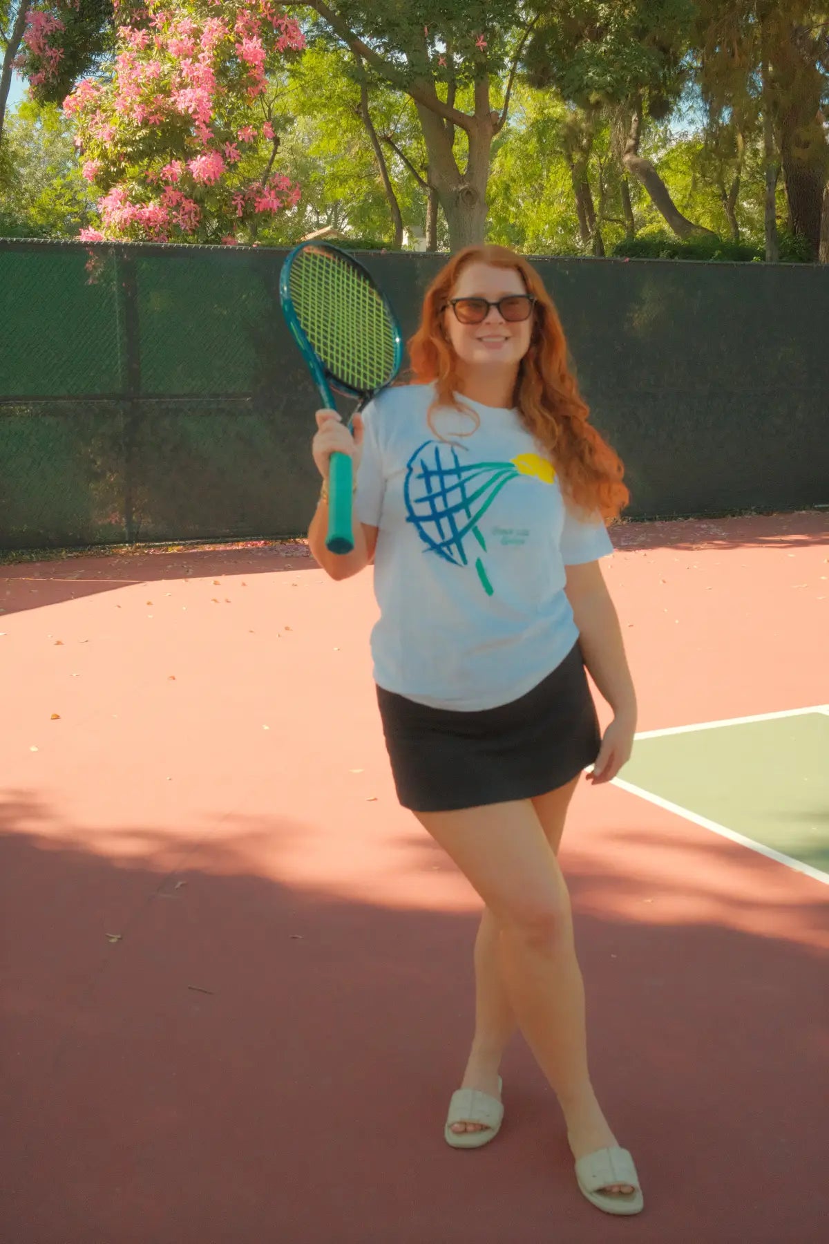 Person with red hair holding a tennis racket on a tennis court with trees and flowers in the background