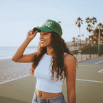 Woman wearing a green tennis trucker hat with 'tennis' text on a beachside court.