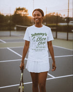 Woman on a tennis court wearing a white t-shirt with text and a skirt, holding a tennis racket.