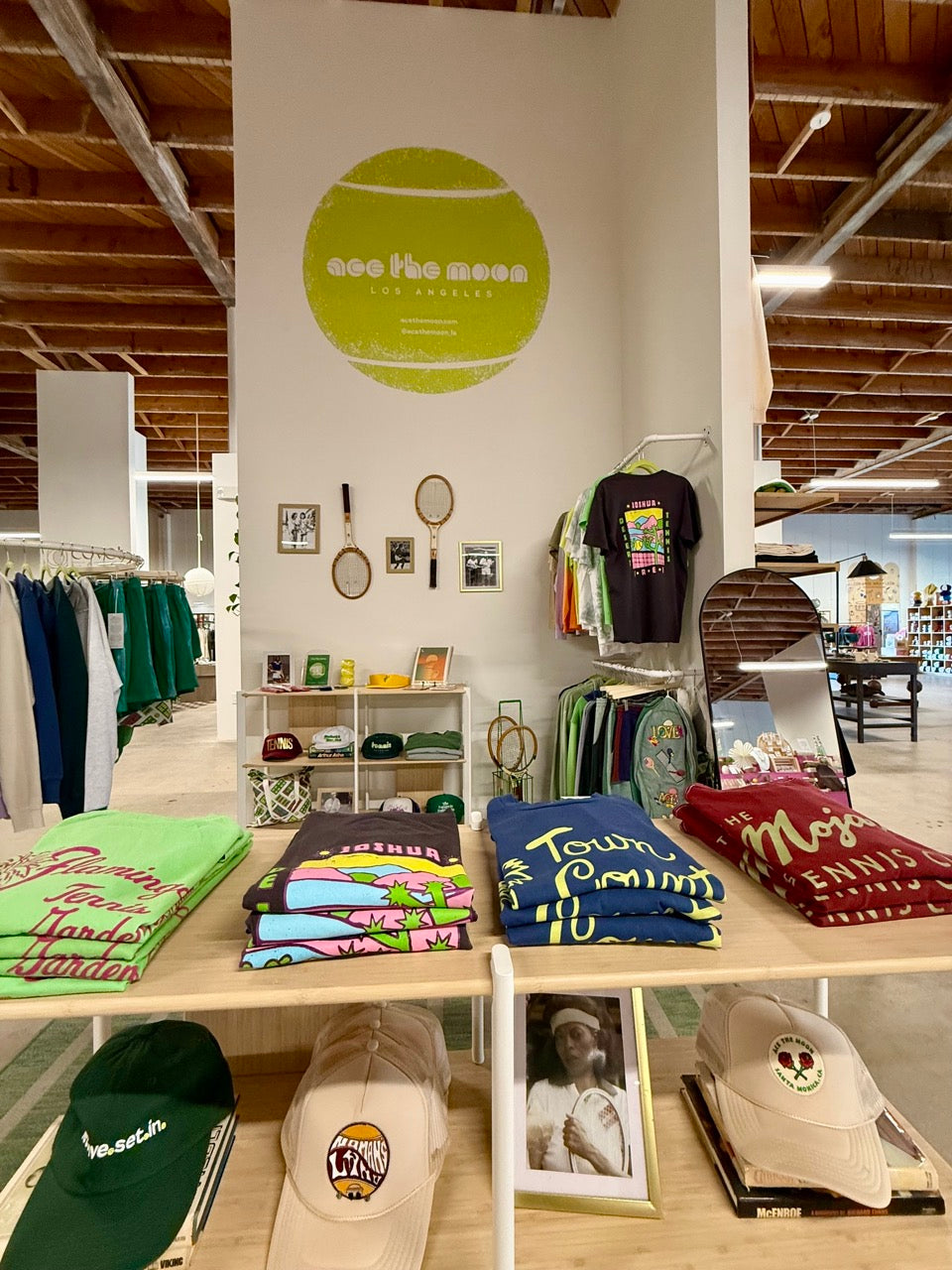 A retail display of 'Ace The Moon' brand merchandise, featuring folded graphic t-shirts, colorful towels, and baseball caps on wooden tables inside a store with a visible 'see the moon' logo on the wall.
