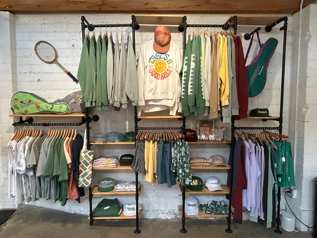 An Ace the Moon retail clothing display featuring multiple black metal clothing racks and wooden shelves filled with assorted apparel, including sweatshirts, t-shirts, shorts, and caps. A tennis racket hangs on the left wall, and a green tennis racket case is displayed on a shelf.