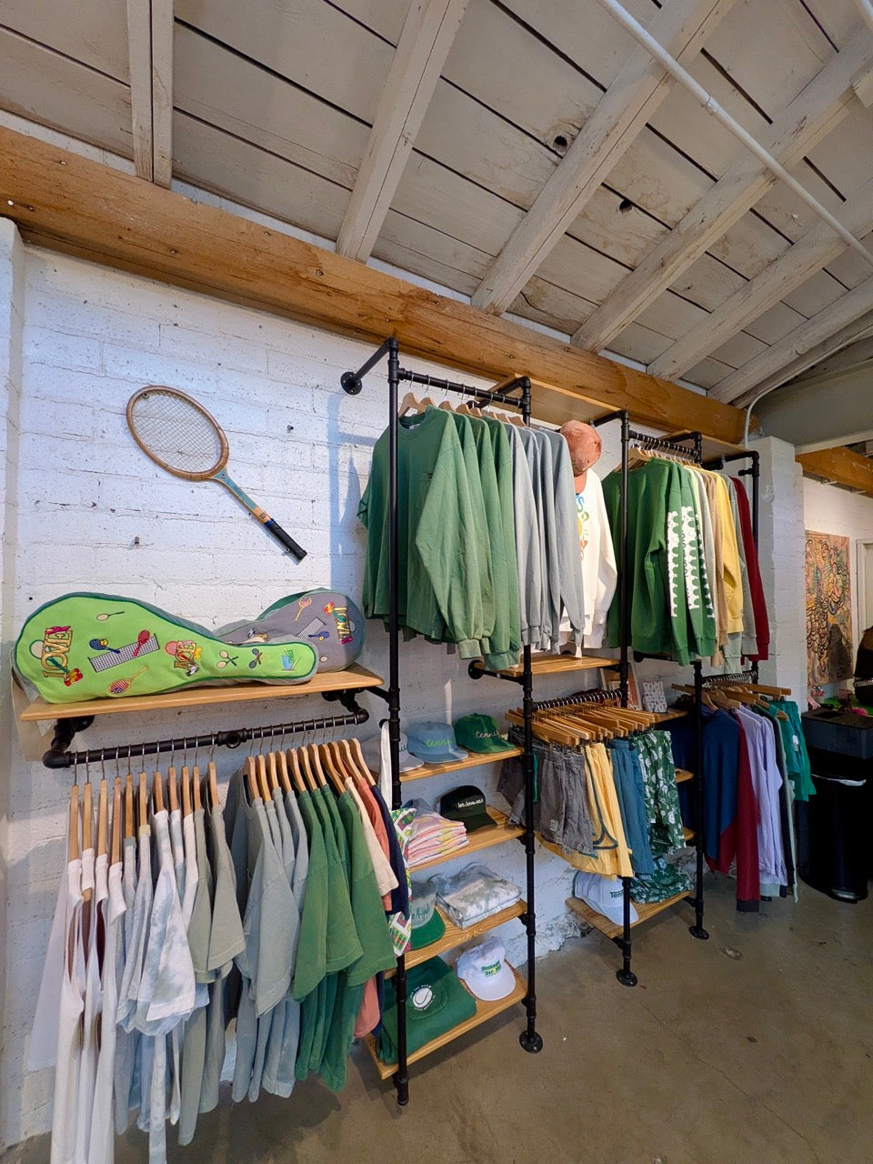 An ace the moon clothing display in a boutique with an exposed wood-beam ceiling and white brick wall. Multiple black clothing racks hold green, gray, white, yellow, and blue garments, with shelves and a wall-mounted racket prop.