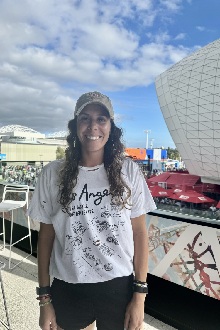 Los Angeles Tennis Bagel tour tee shirt in white by Ace the Moon Premium athleisure apparel brand. shirt worn by a woman standing in front of a tennis stadium