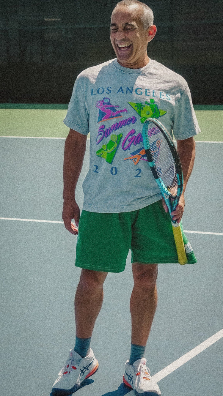Man holding a tennis racket on a tennis court wearing a colorful tennis shirt that says los angeles summer games 2025 by ace the moon and green shorts.