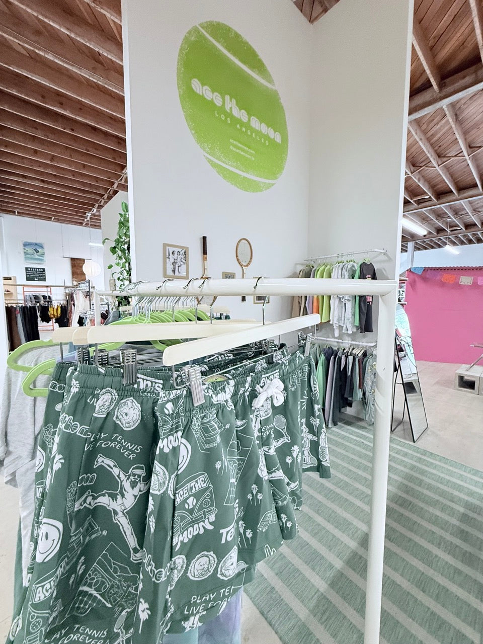 Green patterned athletic shorts by ace the moon hanging on a white clothing rack inside a retail store, with other tennis garments and store fixtures visible in the background.