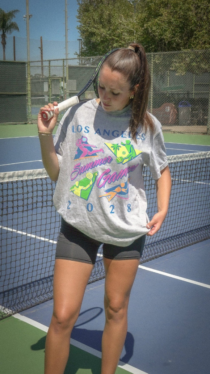 Woman on a tennis court holding a racket, wearing a colorful 'Los Angeles Summer Games' t-shirt.