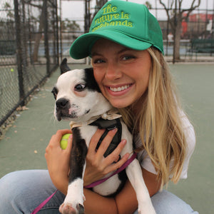 the future is forehands tennis trucker hat in green by Ace the Moon tennis apparel brand LA worn by a girl holding a cute puppy on a tennis court