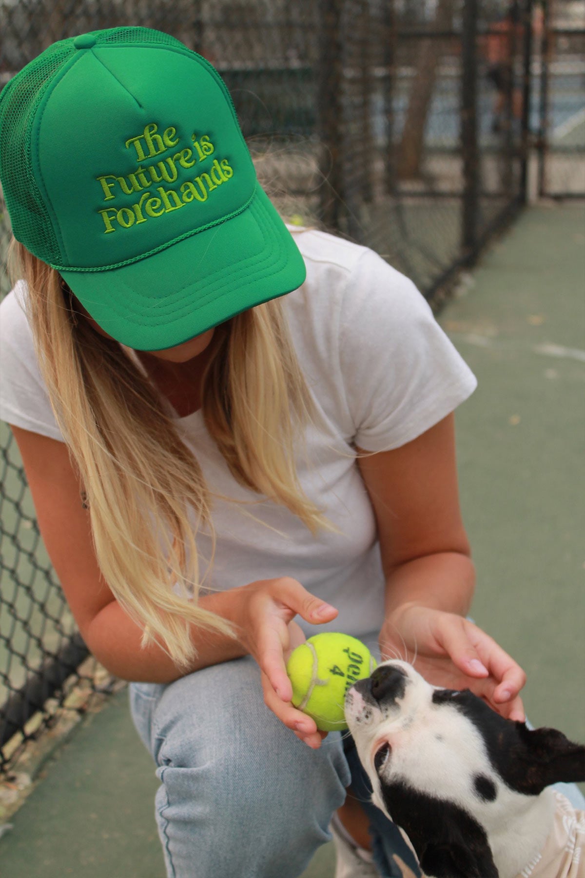 the future is forehands tennis trucker hat in green by Ace the Moon tennis apparel brand LA worn by a blonde girl playing with a puppy and a tennis ball on a tennis court