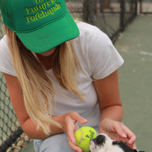 the future is forehands tennis trucker hat in green by Ace the Moon tennis apparel brand LA worn by a blonde girl playing with a puppy and a tennis ball on a tennis court