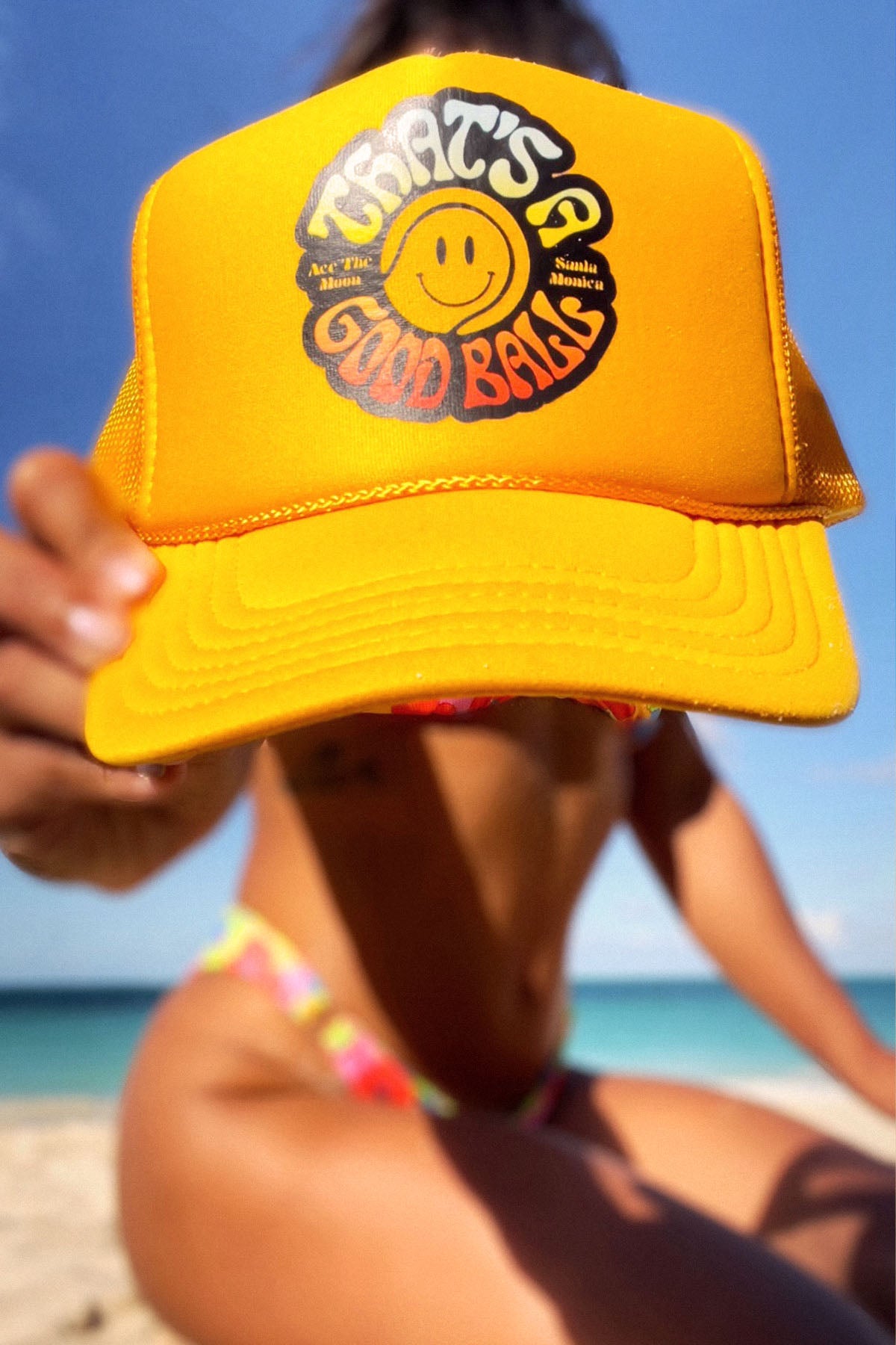 That's A Good Ball Tennis trucker hat in yellow by Ace the Moon LA tennis apparel brand. Female model in bathing suite by the beach holding out the hat 
