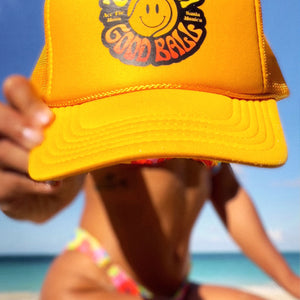 That's A Good Ball Tennis trucker hat in yellow by Ace the Moon LA tennis apparel brand. Female model in bathing suite by the beach holding out the hat 