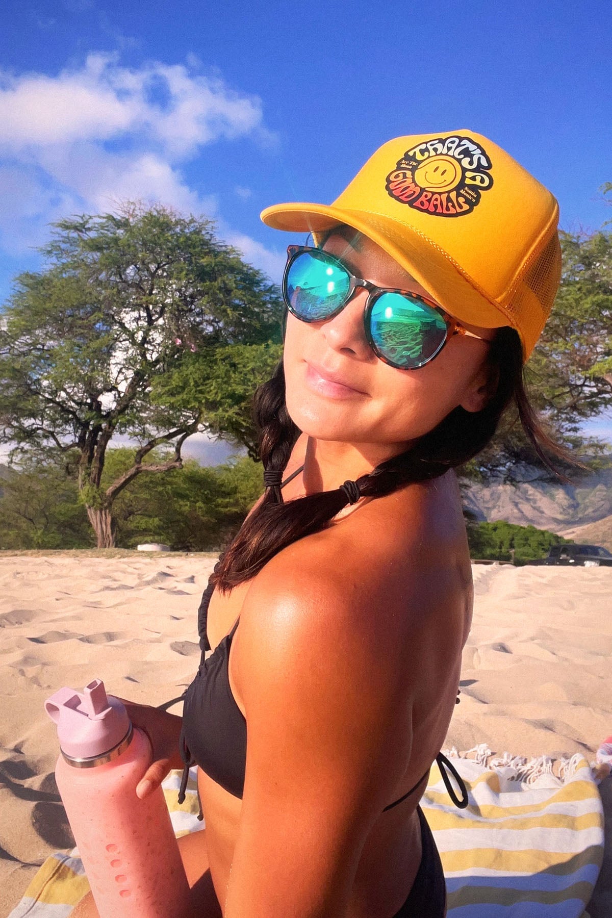That's A Good Ball Tennis trucker hat in yellow by Ace the Moon LA tennis apparel brand. Female model in bathing suite by the beach wearing the hat 
