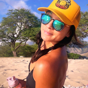 That's A Good Ball Tennis trucker hat in yellow by Ace the Moon LA tennis apparel brand. Female model in bathing suite by the beach wearing the hat 