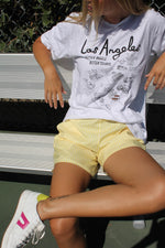 Los Angeles Tennis Bagel tour tee shirt in white by Ace the Moon Premium athleisure apparel brand. shirt worn by a model sitting on a bench on a tennis court
