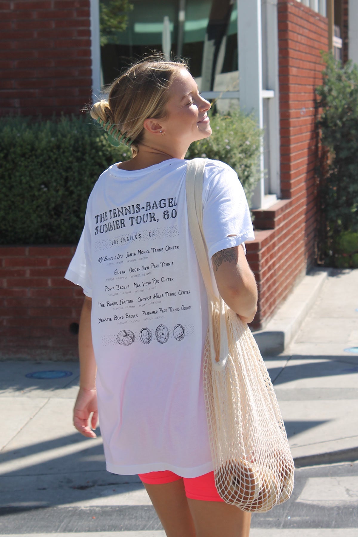 Los Angeles Tennis Bagel tour tee shirt in white by Ace the Moon Premium athleisure apparel brand. shirt worn by a blonde woman walking on a street