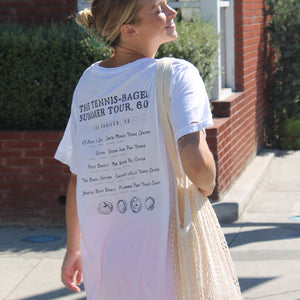Los Angeles Tennis Bagel tour tee shirt in white by Ace the Moon Premium athleisure apparel brand. shirt worn by a blonde woman walking on a street