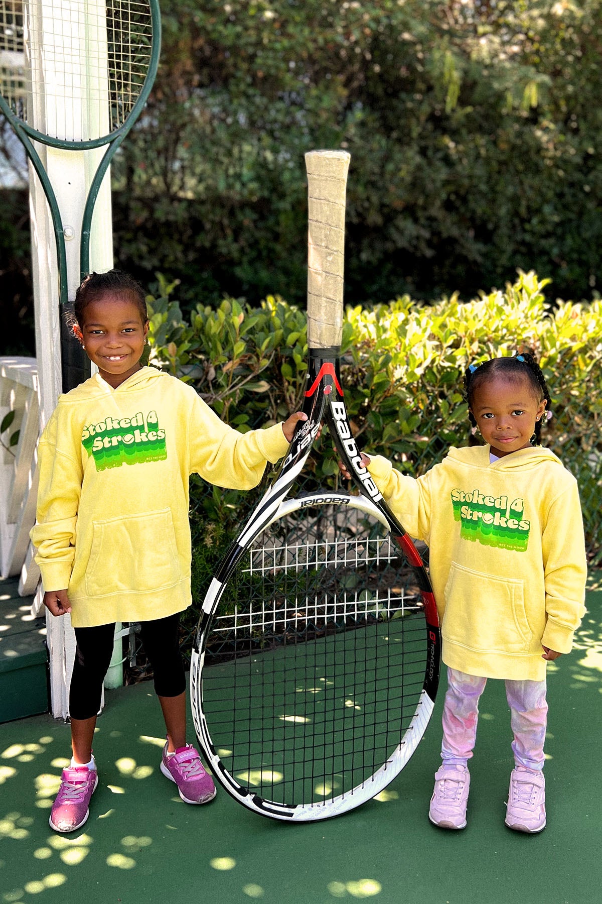 stoked 4 strokes kids hoodie in yellow worn by two adorable kids holding a large oversized tennis racket. sweatshirt made by Ace the Moon LA premium tennis apparel brand